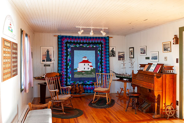 An interior view of a small local museum containing rocking chairs, a piano, a model sailing ship, and a number of framed photographs.